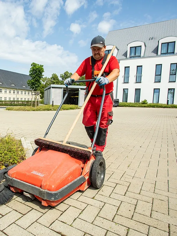 Mitarbeiter schiebt eine Kehrmaschine über einen gepflasterten Innenhof
