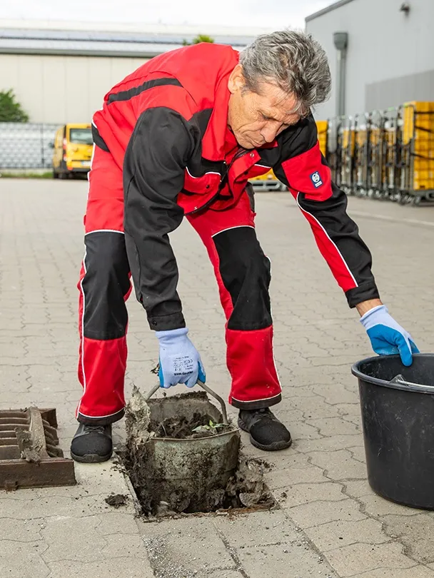 Mitarbeiter hebt den Schlammeimer eines Hofablaufs aus dem Pflaster und leert ihn in einen Eimer